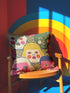 A square decorative pillow cover featuring a faux embroidery pattern of five stylized, expressive doll faces. The central face has yellow hair and pink cheeks. It is surrounded by four other faces in shades of pale beige, green, and bright pink, all with wide staring eyes and open mouths. The design uses intricate, colorful line work to create the illusion of thick woven stitching, but the fabric is a smooth print. Medallions fill the gaps.