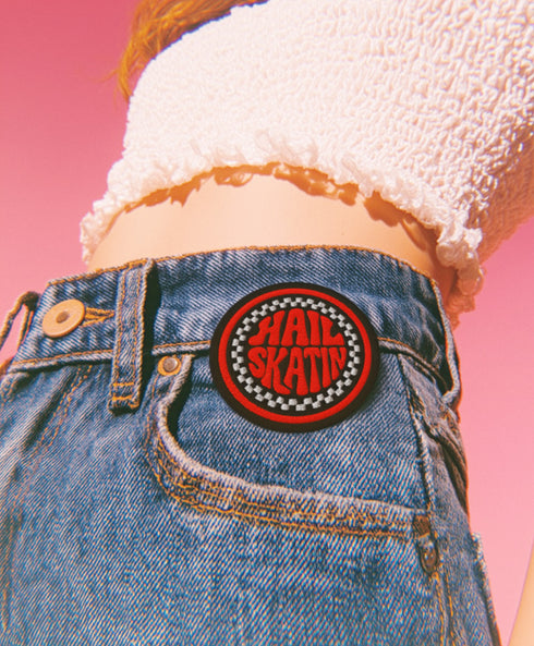 A round, black embroidered patch with a thick, textured merrow border stitched in alternating orange and yellow thread. In the center, the words "PAPERBACK WITCH" are embroidered in a retro 70s-style font. The word "PAPERBACK" is arched across the top in bold orange thread with a thin yellow outline. The word "WITCH" is across the bottom in puffy, bright white thread with a thin yellow outline. Between the words is a central icon of an open book, containing a stylized all-seeing eye.