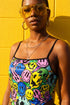 A sleeveless women's camisole top featuring a pointed handkerchief hemline often called a bandana top style. The fabric displays a vibrant all-over psychedelic print of distorting and melting smiley faces mixed with melting skulls. The color palette includes bright neon yellow, electric blue, hot pink, and lime green against a multi-colored background. The top features thin black adjustable spaghetti straps and black trim along the straight neckline. The fit is cropped and form-fitting.