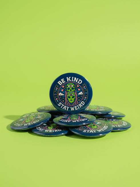 A round 1.75 inch pinback button featuring a retro psychedelic design on a dark navy blue background. The text reads BE KIND STAY WEIRD in vintage cream lettering around the border. The center illustration depicts a neon green skull stacked on top of a neon green alien head. Both the skull and alien have opening third eyes on their foreheads. Pink wavy lines frame the faces. The background is decorated with small colorful mushrooms, hearts, stars, and clouds. Glossy finish.