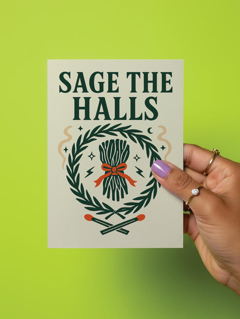 A detailed folk-art style illustration of a "Sage the Halls" holiday greeting card. The design features a bundle of sage smudge sticks tied with a red ribbon at the center of a dark green wreath. The wreath is accented with small crescent moons, stars, and smoke swirls. Below the wreath are two crossed matches with red tips. The card is printed on creamy beige paper with bold, dark green serif text. This funny spiritual and witchy Yule card is a unique pagan greeting for energy cleansing.