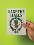 A detailed folk-art style illustration of a "Sage the Halls" holiday greeting card. The design features a bundle of sage smudge sticks tied with a red ribbon at the center of a dark green wreath. The wreath is accented with small crescent moons, stars, and smoke swirls. Below the wreath are two crossed matches with red tips. The card is printed on creamy beige paper with bold, dark green serif text. This funny spiritual and witchy Yule card is a unique pagan greeting for energy cleansing.