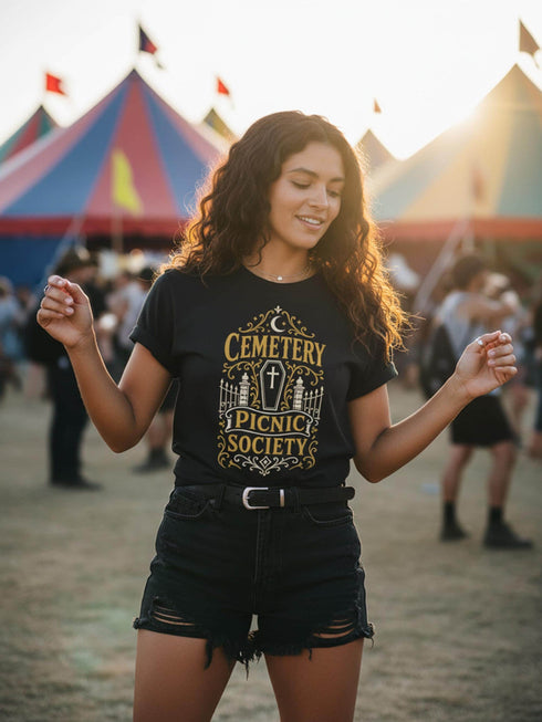 A black unisex crewneck graphic t-shirt featuring an ornate Victorian-inspired emblem printed on the chest in vintage gold and off-white. The centralized, symmetrical design showcases a classic coffin with a cross on the lid, flanked by wrought-iron cemetery gates. Beautiful scrollwork and decorative filigree flourishes surround the bold, retro serif text reading CEMETERY PICNIC SOCIETY. A crescent moon and stars sit at the top, completing this gothic memento mori graphic shirt.