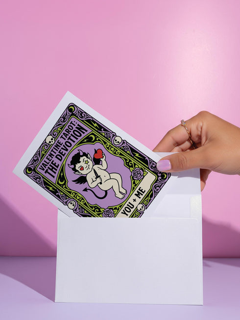 A vertical folded greeting card featuring a pastel goth tarot design titled The Devotion. The central illustration shows a pale devil imp with black wings and horns, holding a red heart in the left hand and a black dagger in the right. The imp has one star eye and one heart eye. The background is lavender purple. A black border frames the scene with teal vines, skulls, gold crescent moons, and roses. The bottom banner reads YOU + ME. The finish is shimmering pearlescent.
