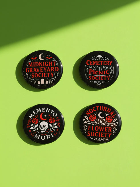 A collection of four glossy black circular pinback buttons featuring striking red and white gothic illustrations. One button shows "Midnight Graveyard Society" text with bats and tombstones. Another reads "Cemetery Picnic Society" with a coffin and umbrella icon. A third features a detailed white skull and red roses with "Memento Mori" text below. The final pin displays "Nocturnal Flower Society" surrounded by floral vines.