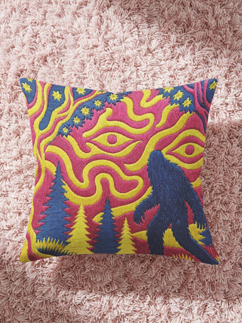 A square decorative throw pillow cover featuring a trippy retro Bigfoot scene. A large navy blue silhouette of Sasquatch walks through a forest of navy and yellow pine trees. The background is a psychedelic swirling sky in vibrant pink and yellow, featuring a large stylized eye and a cluster of small yellow stars. The flat two dimensional print visually mimics the textured look of heavy needlepoint embroidery. The pillow features a sleek hidden zipper on the bottom. No insert is included.