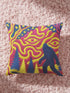 A square decorative throw pillow cover featuring a trippy retro Bigfoot scene. A large navy blue silhouette of Sasquatch walks through a forest of navy and yellow pine trees. The background is a psychedelic swirling sky in vibrant pink and yellow, featuring a large stylized eye and a cluster of small yellow stars. The flat two dimensional print visually mimics the textured look of heavy needlepoint embroidery. The pillow features a sleek hidden zipper on the bottom. No insert is included.