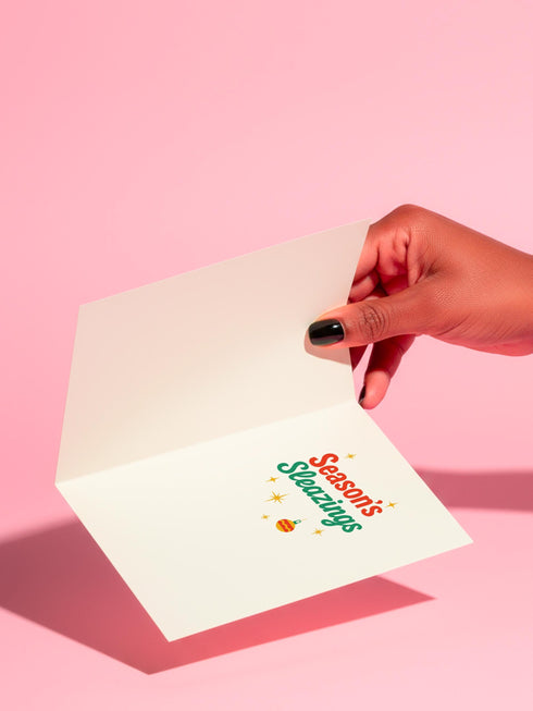 A 5x7 pearlescent holiday card by HoneyDazed. The card illustration, inspired by John Waters, features the drag queen Divine in a red dress with a Santa hat. She points a gun, withtext reads "Have Yourself a Filthy Christmas" in campy font. The card is bordered by garland. Iinside of the card reads: "Season's Sleazings" in red and green text. This kitschy greeting card is a perfect piece of cult film memorabilia and a funny adult humor gift for fans of queer art.