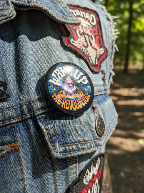 A round pinback button featuring a retro psychedelic design. The central focus is a stylized Eye of Providence in a pink triangle, surrounded by vibrant orange and yellow flames. Behind the triangle are teal and petrol blue sunburst rays. The text "WAKE UP" curves across the top in cream wavy letters, and "THE REVOLUTION" curves along the bottom in orange. The button has a glossy finish and is available in 1.75 inch or 2.25 inch sizes.