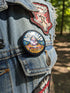 A round pinback button featuring a retro psychedelic design. The central focus is a stylized Eye of Providence in a pink triangle, surrounded by vibrant orange and yellow flames. Behind the triangle are teal and petrol blue sunburst rays. The text "WAKE UP" curves across the top in cream wavy letters, and "THE REVOLUTION" curves along the bottom in orange. The button has a glossy finish and is available in 1.75 inch or 2.25 inch sizes.