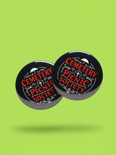 A collection of four glossy black circular pinback buttons featuring striking red and white gothic illustrations. One button shows "Midnight Graveyard Society" text with bats and tombstones. Another reads "Cemetery Picnic Society" with a coffin and umbrella icon. A third features a detailed white skull and red roses with "Memento Mori" text below. The final pin displays "Nocturnal Flower Society" surrounded by floral vines.