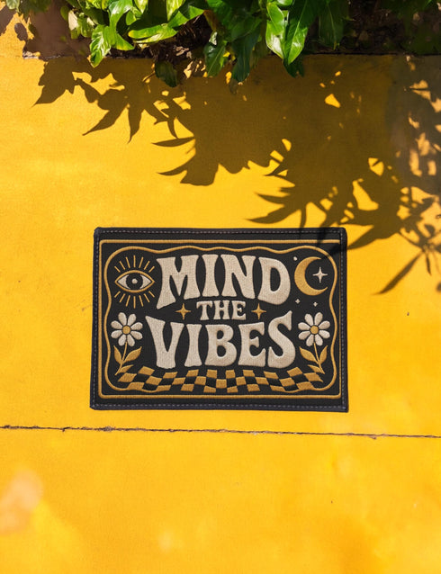 A rectangular floor mat with a dark charcoal black background. The center features the phrase 'MIND THE VIBES' in stylized, wavy cream and golden-mustard retro typography. The text is surrounded by symmetrical motifs: a golden evil eye with sun rays on the top left, a golden crescent moon with a four-point star on the top right, and two cream daisy flowers with golden stems at the bottom corners. The bottom edge has a wavy golden checkerboard pattern. A faux-stitch border frames the art.