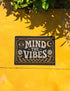 A rectangular floor mat with a dark charcoal black background. The center features the phrase 'MIND THE VIBES' in stylized, wavy cream and golden-mustard retro typography. The text is surrounded by symmetrical motifs: a golden evil eye with sun rays on the top left, a golden crescent moon with a four-point star on the top right, and two cream daisy flowers with golden stems at the bottom corners. The bottom edge has a wavy golden checkerboard pattern. A faux-stitch border frames the art.