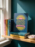 A hardcover spiral planner for 2027 with a dark blue cover and retro 70s style graphics. The title reads "2027 SHITSHOW All-Seeing Agenda" in bold yellow and pink text. Below the title is a large, stylized all-seeing eye with pink rays emanating from it. At the bottom, a pink ribbon contains the text "BLINK TWICE FOR MERCY." The back cover is also dark blue with a yellow sun graphic and the text "Plans bloom. Deadlines hum. I write, I wander, then get it done. Keep receipts. Keep grace.