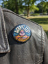 A round pinback button featuring a retro psychedelic design. The central focus is a stylized Eye of Providence in a pink triangle, surrounded by vibrant orange and yellow flames. Behind the triangle are teal and petrol blue sunburst rays. The text "WAKE UP" curves across the top in cream wavy letters, and "THE REVOLUTION" curves along the bottom in orange. The button has a glossy finish and is available in 1.75 inch or 2.25 inch sizes.
