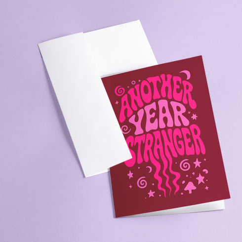 A pink greeting card with the text "Another Year Stranger" printed on it, placed on a purple background.