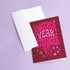 A pink greeting card with the text "Another Year Stranger" printed on it, placed on a purple background.