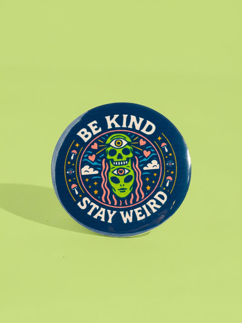 A round 1.75 inch pinback button featuring a retro psychedelic design on a dark navy blue background. The text reads BE KIND STAY WEIRD in vintage cream lettering around the border. The center illustration depicts a neon green skull stacked on top of a neon green alien head. Both the skull and alien have opening third eyes on their foreheads. Pink wavy lines frame the faces. The background is decorated with small colorful mushrooms, hearts, stars, and clouds. Glossy finish.