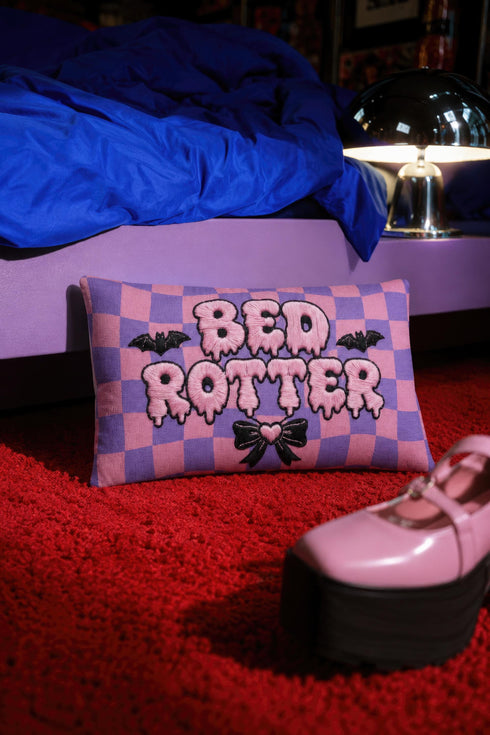 Pastel goth lumbar pillow, 20 by 12 inches, with a pink and lavender purple wavy checkerboard background. The words Bed Rotter appear in dripping pink bubble letters with a faux embroidery thread texture and black outlines. Two black bats with pink stitch details flank the text, and a black bow with a pink heart center sits below. Double sided flat print that mimics raised embroidery on linen feel polyester with a hidden zipper, insert included.