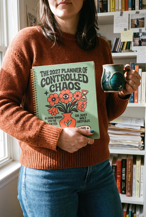 A sage green hardcover spiral planner for 2027-2028 with the title "The 2027 Planner of Controlled Chaos" in a funky, retro 70s-style font. The cover features a groovy illustration of orange and pink flowers in a vase, with one flower depicted as an eyeball. The text reads "Bloom where you're planted. Or just wilt artfully." and "Notes from the Void." The planner has a gold metal wire-o binding. The back cover shows a decorative floral frame with a "Property Of" section
