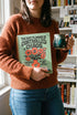 A sage green hardcover spiral planner for 2027-2028 with the title "The 2027 Planner of Controlled Chaos" in a funky, retro 70s-style font. The cover features a groovy illustration of orange and pink flowers in a vase, with one flower depicted as an eyeball. The text reads "Bloom where you're planted. Or just wilt artfully." and "Notes from the Void." The planner has a gold metal wire-o binding. The back cover shows a decorative floral frame with a "Property Of" section