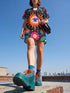 A short-sleeve, above-the-knee t-shirt dress with a straight, structured fit. The dress has a true black background featuring a bold, vibrant graphic pattern. The center displays a large blue and white evil eye surrounded by a bright orange wavy sunburst with a white jagged border. Above and below the eye are bright pink daisy flowers with orange centers, scattered white four-petal stars, and wavy blue and teal green leaf shapes. The fabric is thick, smooth, and holds its clean shape.