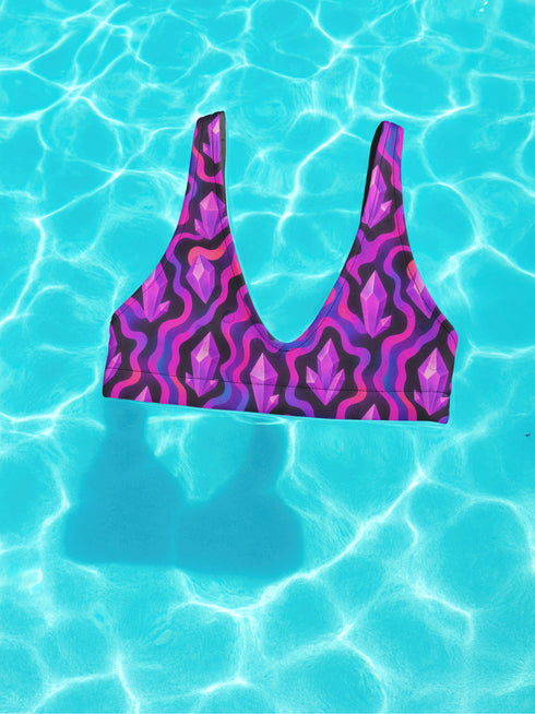 Psychedelic crystal bikini top featuring a vivid print of pink and purple crystals with wavy neon accents on a black background. Sports bra-style top with a scoop neckline, wide shoulder straps, and removable padding. Made of recycled polyester and LYCRA XTRALIFE with UPF 50+ sun protection. Available in sizes XS to 3XL. Double-layered construction with a snug, secure fit. Sustainable swimwear top for rave, beach, or summer festival wear.