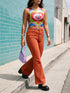 A sleeveless scoop neck crop top featuring a bold all-over printed design. The central graphic is a large bright red heart enclosing a scalloped pink flower shape and a blue evil eye motif in the middle. The background surrounds the heart with mirrored concentric wavy arches and scalloped borders in vibrant shades of lime green, pink, blue, and red. The fitted garment has a smooth athletic texture. Available in a standard matte performance knit or a sparkly silver glitter foil finish.
