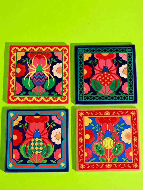 A set of four square coasters featuring a glossy glazed finish and vibrant insect illustrations. Each coaster measures approximately four by four inches. The designs showcase symmetrical beetles and spiders in bold red, blue, and orange colors, framed by intricate floral and geometric borders in a maximalist folk art style. Available in ceramic with cork backing or hardboard MDF. The edges are smooth. The designs are vivid and highly saturated, designed to provide grip and protect furniture.