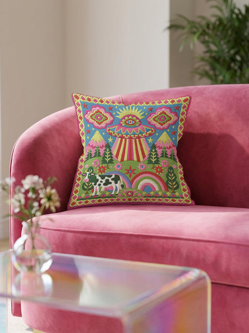 Description of a square throw pillow with a vibrant maximalist alien design. A central UFO with a large all-seeing eye hovers above a landscape with two rainbows, pink mountains, and green pine trees. A black and white spotted cow stands in the foreground. The design is a high-contrast palette of sky blue, magenta, hot pink, and meadow green. The pillow has a faux-embroidery 2D print finish on linen-feel fabric, framed by a yellow and pink scalloped floral border.