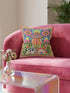 Description of a square throw pillow with a vibrant maximalist alien design. A central UFO with a large all-seeing eye hovers above a landscape with two rainbows, pink mountains, and green pine trees. A black and white spotted cow stands in the foreground. The design is a high-contrast palette of sky blue, magenta, hot pink, and meadow green. The pillow has a faux-embroidery 2D print finish on linen-feel fabric, framed by a yellow and pink scalloped floral border.