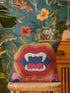 A colorful, patterned throw pillow with a graphic of a pair of open, fanged lips is sitting on a wicker chair.