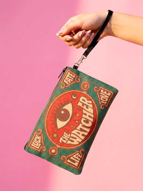 A rectangular 9x6 inch makeup bag and zipper pouch featuring a textured linen-like polyester fabric. The bag showcases a bold, maximalist print with vivid colors typical of retro 70s and pastel goth aesthetics, centering on a large mystic eye design labeled "The Watcher." It features a sturdy black zipper across the top and includes a removable black nylon wristlet strap.Perfect for use as a pencil case, cosmetic organizer, or festival wristlet.