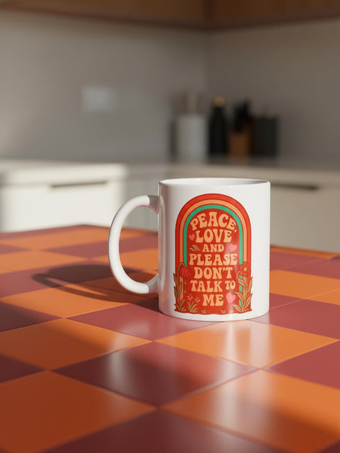 A glossy white porcelain ceramic mug featuring a vibrant maximalist design centered on the exterior. The artwork displays a retro 1970s-inspired rainbow arch composed of concentric bands in burnt orange, mustard yellow, and teal green. Inside the arch, stylized vintage bubble-serif typography in reddish-orange reads PEACE LOVE AND PLEASE DON'T TALK TO ME. The design evokes a groovy, hippie aesthetic mixed with sassy introvert humor.