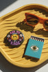 A round glossy metal pinback button featuring a flat 2D print design on a solid black background. In the center is an angry yellow smiley face surrounded by orange and red flames. Above the smiley face, pink text reads ASK ME and white text reads ABOUT. Below the face, white text reads MY and large purple text reads RAGE. The design is accented with two white daisies, two purple flowers, pink and orange four-point stars, and orange lightning bolts framing the outer edges.