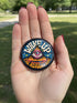 A round pinback button featuring a retro psychedelic design. The central focus is a stylized Eye of Providence in a pink triangle, surrounded by vibrant orange and yellow flames. Behind the triangle are teal and petrol blue sunburst rays. The text "WAKE UP" curves across the top in cream wavy letters, and "THE REVOLUTION" curves along the bottom in orange. The button has a glossy finish and is available in 1.75 inch or 2.25 inch sizes.