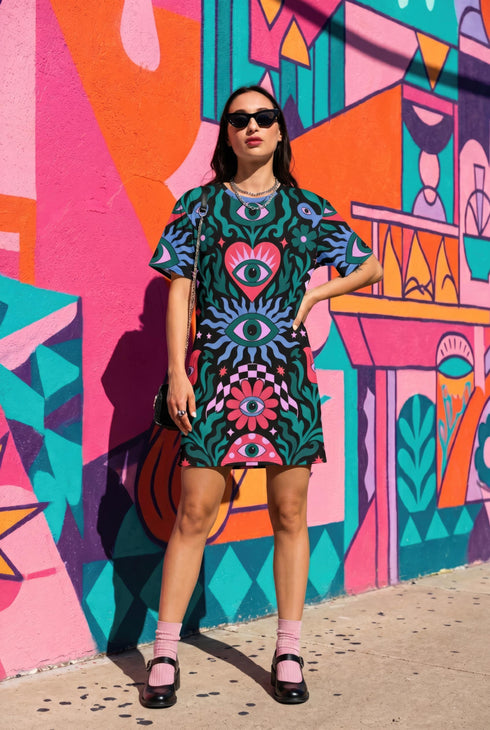 A short-sleeve, above-the-knee t-shirt dress with a true black background. The front features a vibrant, vertically stacked symmetrical graphic design. From top to bottom, the design includes a pink heart containing a green and blue eye, a large blue sunburst with a central eye, a red daisy flower with an eye flanked by pink and black checkered arches, and a pink mushroom cap with an eye. Swirling emerald green foliage, pink stars, and purple stars fill the negative space around the motifs.