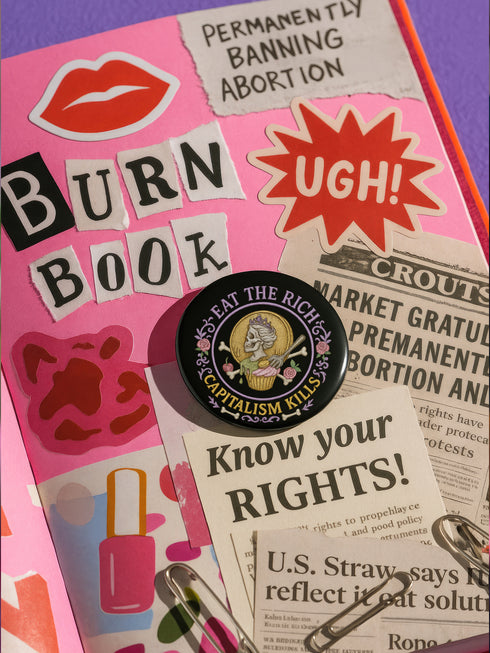 Glossy black pinback button featuring a pastel embroidered-style design of a skeleton queen on a cupcake, surrounded by roses and bones, with the text “Eat the Rich” and “Capitalism Kills.” Displayed on protest props, tea cups, and pastel backgrounds.