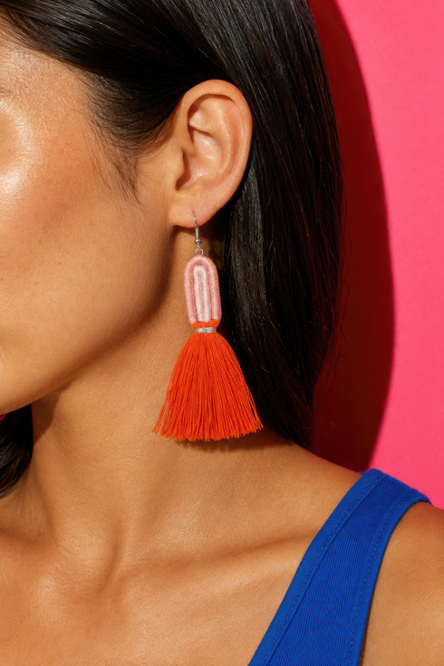 A close-up of a woman's ear wearing a pair of orange tassel earrings with a pink tassel at the end.