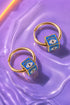 two gold hoop earrings with blue square pendants, each containing an eye design, resting on a purple surface.