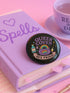 Glossy black pinback button with pastel artwork of a bubbling cauldron, rainbow, potion bottles, and tools. Text reads “Queer Coven, Hex Proud.”