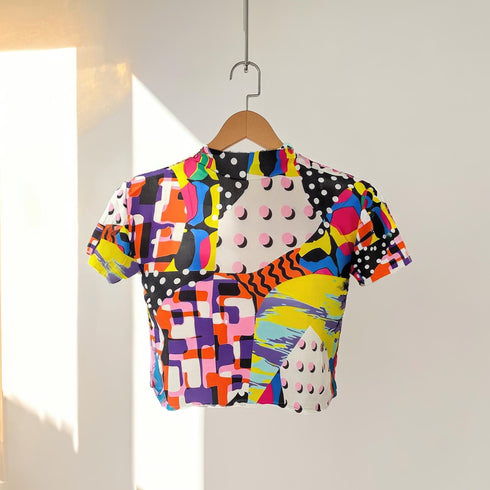 A colorful 90s-style mesh crop top featuring a maximalist abstract pattern. This see-through shirt has a high mock neck, short sleeves, and a vibrant, retro design with geometric shapes, color blocking, and polka dots in bold shades of pink, yellow, blue, and orange. The Y2K-inspired top is a statement piece of festival clothing, perfect for a rave outfit or alternative, dopamine-dressing-inspired streetwear looks.