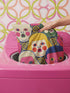 A square decorative pillow cover featuring a faux embroidery pattern of five stylized, expressive doll faces. The central face has yellow hair and pink cheeks. It is surrounded by four other faces in shades of pale beige, green, and bright pink, all with wide staring eyes and open mouths. The design uses intricate, colorful line work to create the illusion of thick woven stitching, but the fabric is a smooth print. Medallions fill the gaps.