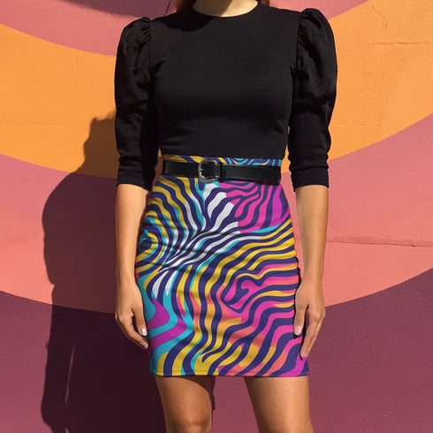 A woman wearing a black top and a colorful, patterned skirt stands in front of a vibrant, multicolored wall.