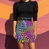 A woman wearing a black top and a colorful, patterned skirt stands in front of a vibrant, multicolored wall.