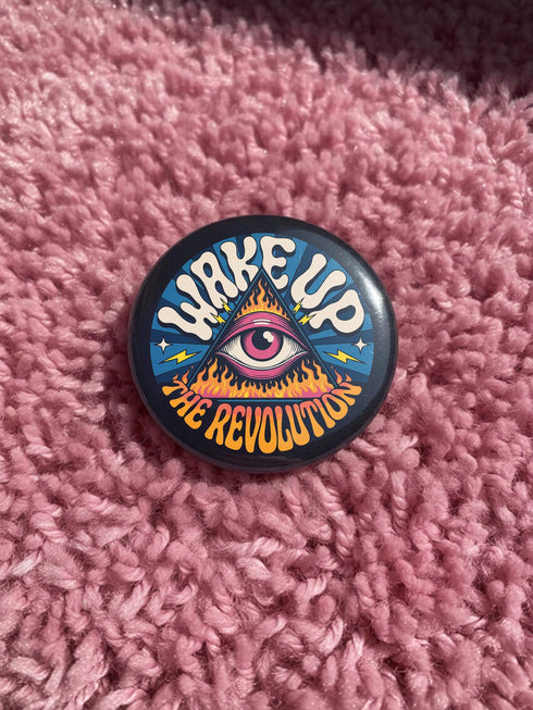 A round pinback button featuring a retro psychedelic design. The central focus is a stylized Eye of Providence in a pink triangle, surrounded by vibrant orange and yellow flames. Behind the triangle are teal and petrol blue sunburst rays. The text "WAKE UP" curves across the top in cream wavy letters, and "THE REVOLUTION" curves along the bottom in orange. The button has a glossy finish and is available in 1.75 inch or 2.25 inch sizes.