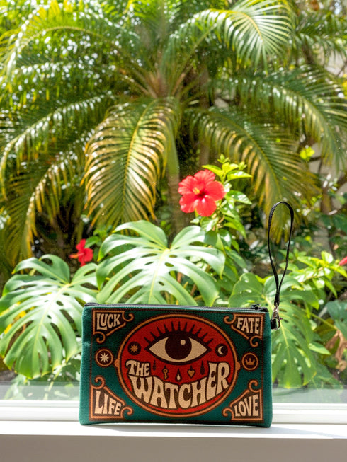 A rectangular 9x6 inch makeup bag and zipper pouch featuring a textured linen-like polyester fabric. The bag showcases a bold, maximalist print with vivid colors typical of retro 70s and pastel goth aesthetics, centering on a large mystic eye design labeled "The Watcher." It features a sturdy black zipper across the top and includes a removable black nylon wristlet strap.Perfect for use as a pencil case, cosmetic organizer, or festival wristlet.