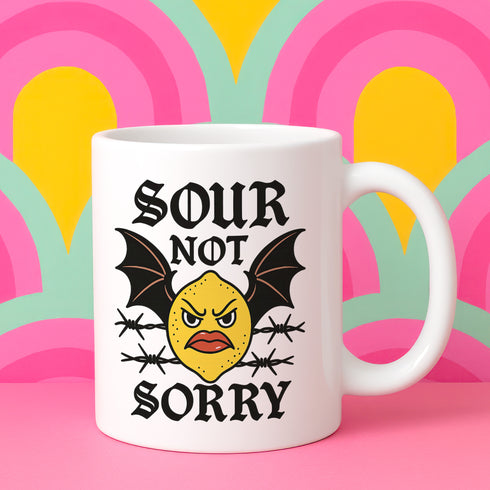 White ceramic 11oz coffee mug featuring a bold tattoo-style graphic with an angry yellow lemon face, black bat wings, and barbed wire details. The design reads “SOUR NOT SORRY” in gothic black lettering above and below the lemon. C-handle, durable construction, dishwasher and microwave safe. Graphic is high-contrast and punk-inspired, perfect for adding attitude to your drinkware collection.