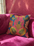 A vibrant, colorful throw pillow with a floral design rests on a pink velvet couch, bathed in soft sunlight streaming through a nearby window.