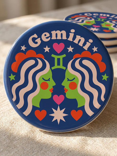 A close-up photograph of a round, glossy ceramic coaster featuring a bold retro pop art design. The background is a deep royal blue. In the center are two symmetrical profile faces of twins looking at each other, featuring bright green skin, closed eyes, and red cheeks. They have wavy white and pale pink hair with red accents. The word Gemini is arched at the top in a retro white font. The design is decorated with a green Gemini symbol, pink and red hearts, and white starbursts.